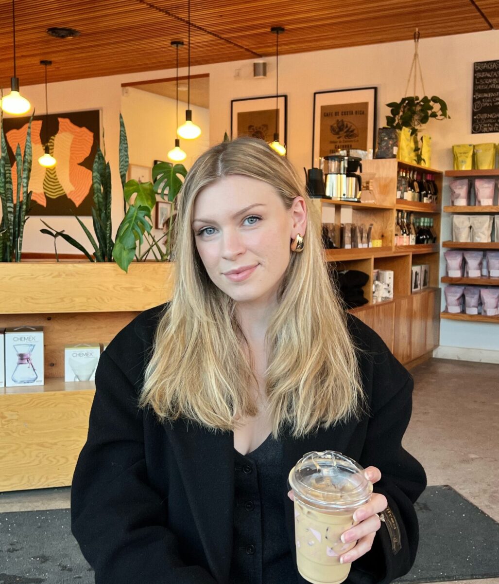 Cream-colored beverage served in a clear cup with a domed lid, held by a young woman, in a cozy café interior.